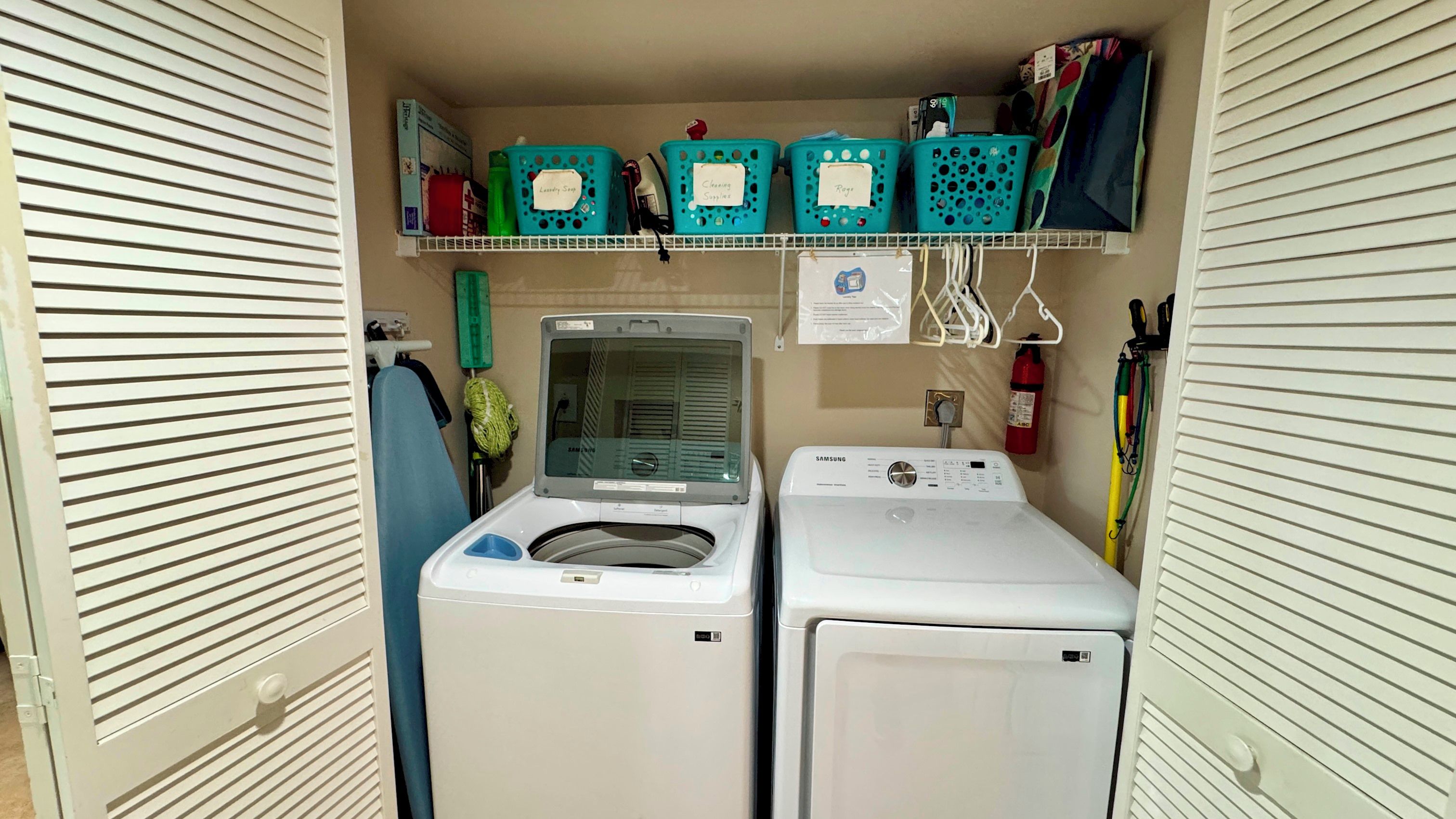 A laundry room with two washing machines, a drying rack, and labeled baskets for laundry supplies.