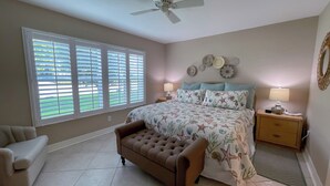 A bedroom with a bed, two bedside tables, a chair, a ceiling fan, and a large window with blinds. - Essex 106N: 5 Min walk to Beach, Movies, Restaurants (Marco Island)