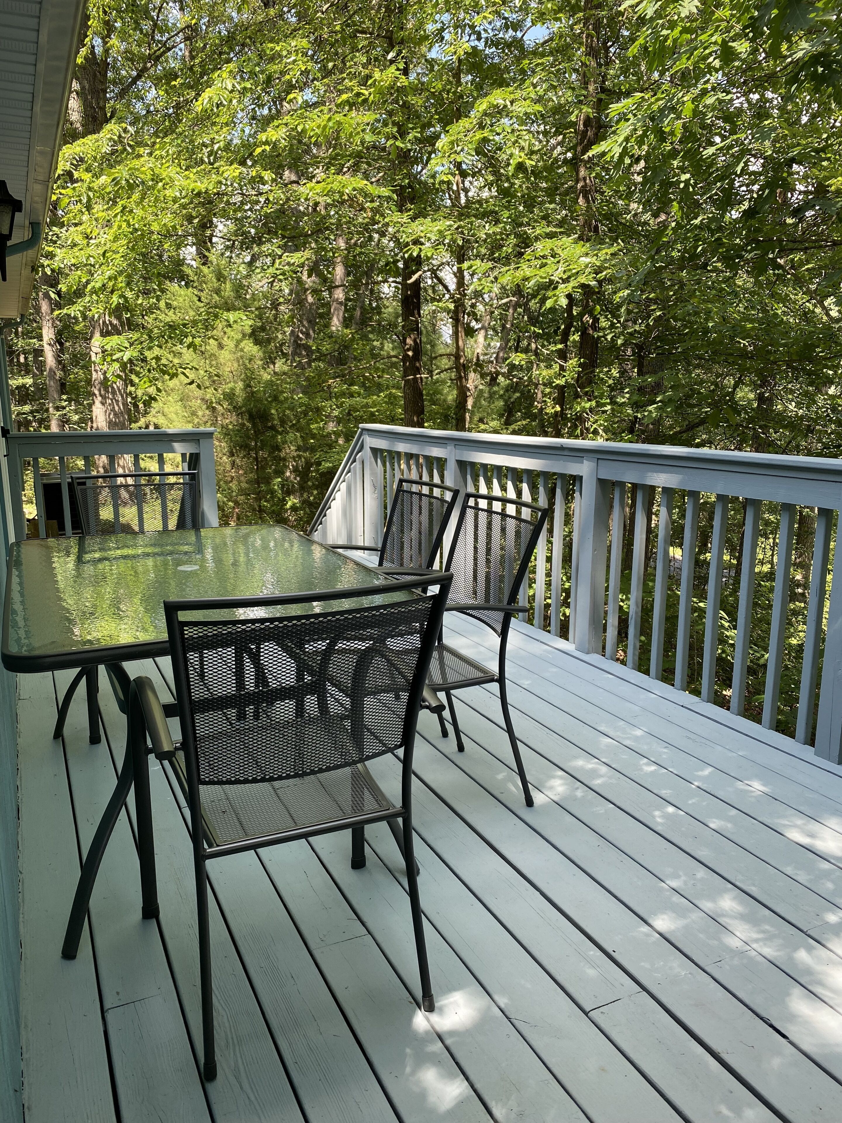 A wooden deck with a glass table and metal chairs, surrounded by trees.