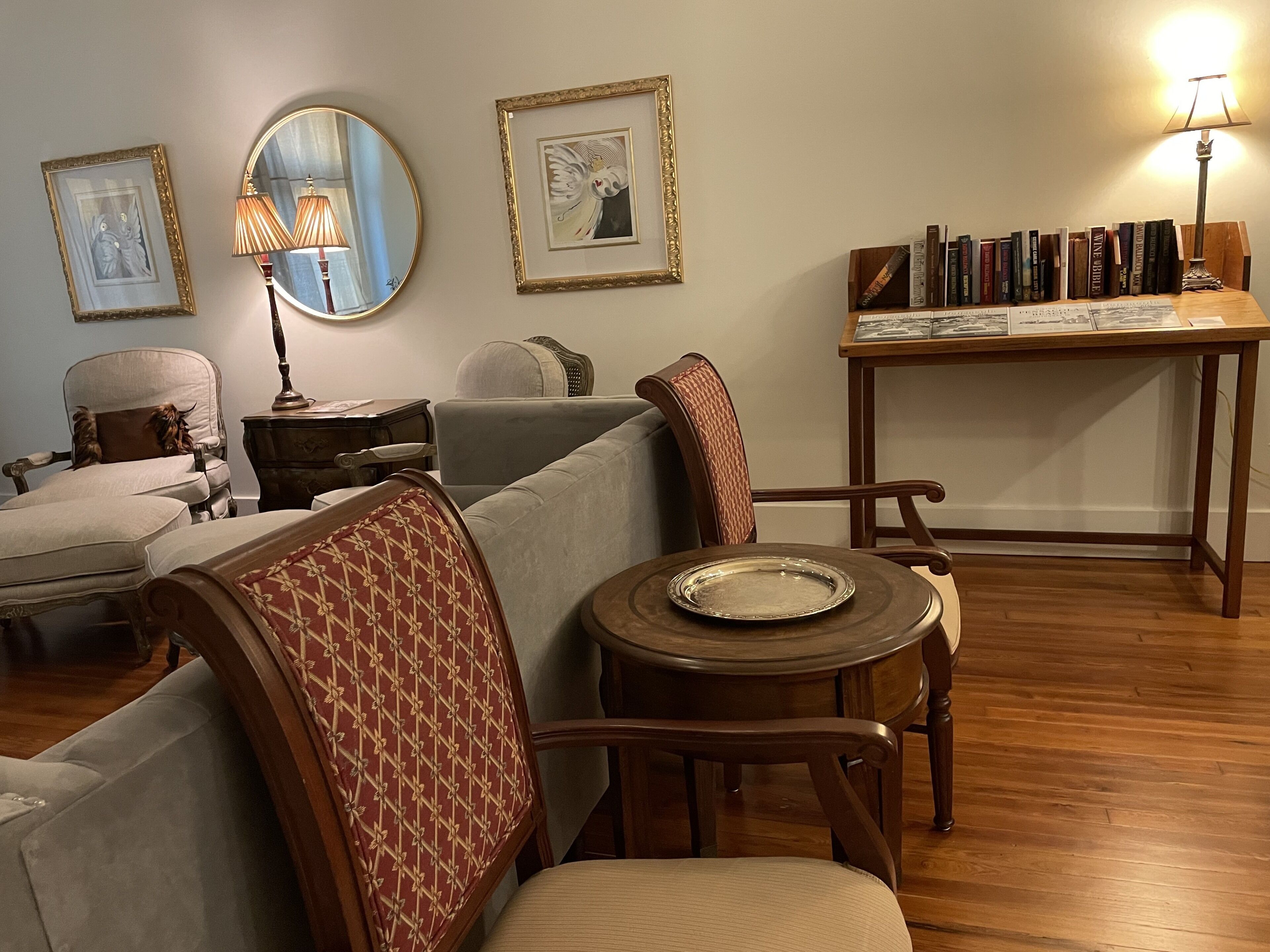 A living room with a sofa, armchairs, a coffee table, a side table, a lamp, framed artwork, and a bookshelf.