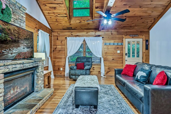 A cozy cabin interior with a stone fireplace, a leather sofa, and a ceiling fan.