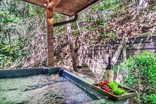 A wine bottle and glasses on a table with grapes and a small dish, set outdoors with a wooden structure overhead and greenery in the background. - Secluded Charming Smokies Cabin Minutes from Attractions (Gatlinburg)