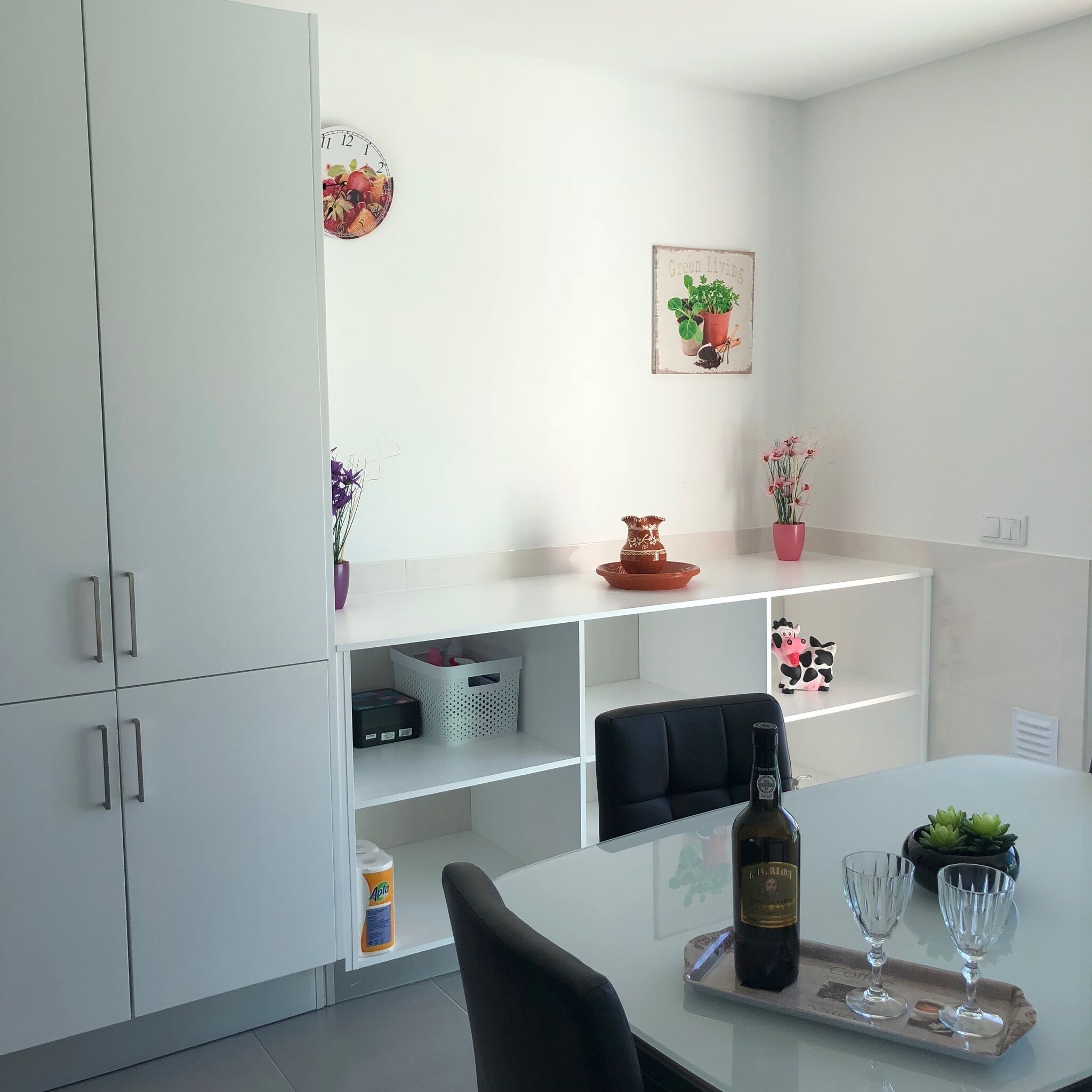 Una cocina moderna con armarios blancos, una mesa de comedor con dos copas de vino, una botella de vino y varios objetos decorativos, incluyendo un reloj, plantas en macetas y un cuadro enmarcado.