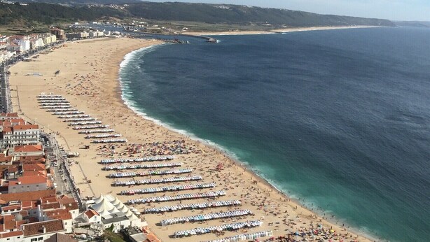 Una spiaggia costiera con file di ombrelloni e lettini, una battigia sabbiosa e edifici lungo la costa.