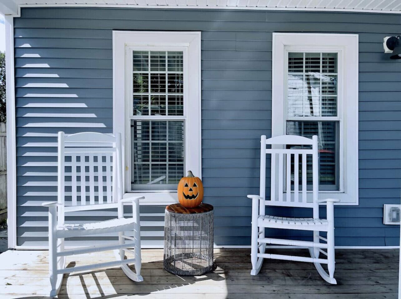 A porch with two white rocking chairs and a metal stool, each with a carved pumpkin.