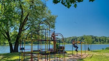 Explore the playground's multiple levels and climbing elements in a serene park setting surrounded by trees and a body of water.