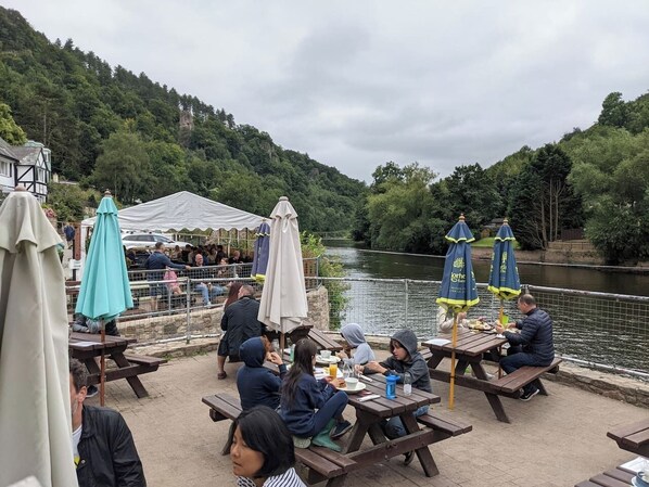An outdoor dining area with picnic tables and umbrellas, overlooking a river. - Casita Lodge | Hot tub Stay for two in the Forest of Dean (Cinderford)