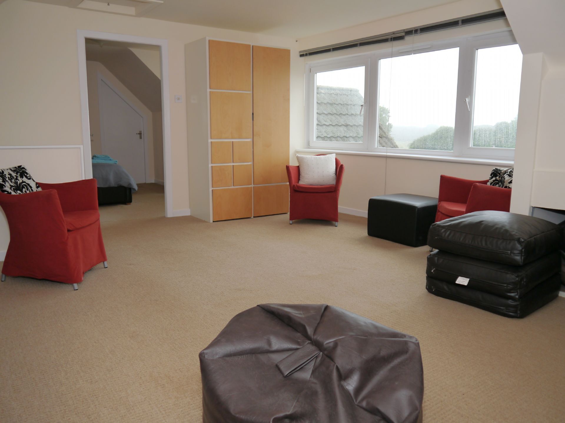 Une pièce avec une grande fenêtre, une armoire, un fauteuil rouge, un pouf noir et un lit recouvert d’une couverture bleue.