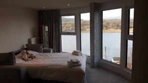 Ein Hotelzimmer mit einem großen Bett, einem Sessel, einem Fenster mit Blick auf einen See und Berge sowie einem Teddybären auf dem Bett.