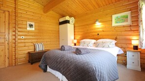 A log cabin bedroom with a bed, bedside tables, a nightstand, a dresser, and a wall-mounted lamp. - Birchlea -  a lodge that sleeps 6 guests  in 3 bedrooms (Kippford, Dalbeattie)