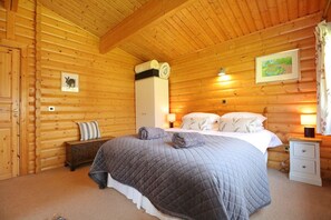 Une chambre à coucher aménagée dans un chalet en rondins, avec un lit, des tables de chevet, une petite table, une commode et une lampe fixée au mur.
