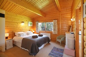 A wooden cabin bedroom with a bed, bedside tables, a chair, a window, and a dresser. - Birchlea -  a lodge that sleeps 6 guests  in 3 bedrooms (Kippford, Dalbeattie)