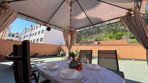 An outdoor dining area with a table set for four, under a large umbrella, with potted plants and a view of buildings and trees.