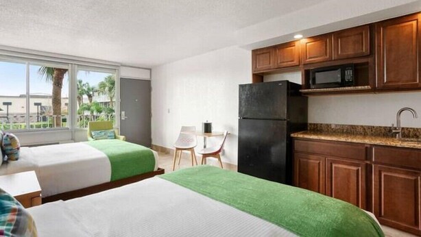A hotel room with two beds, a kitchenette, and a view of palm trees.