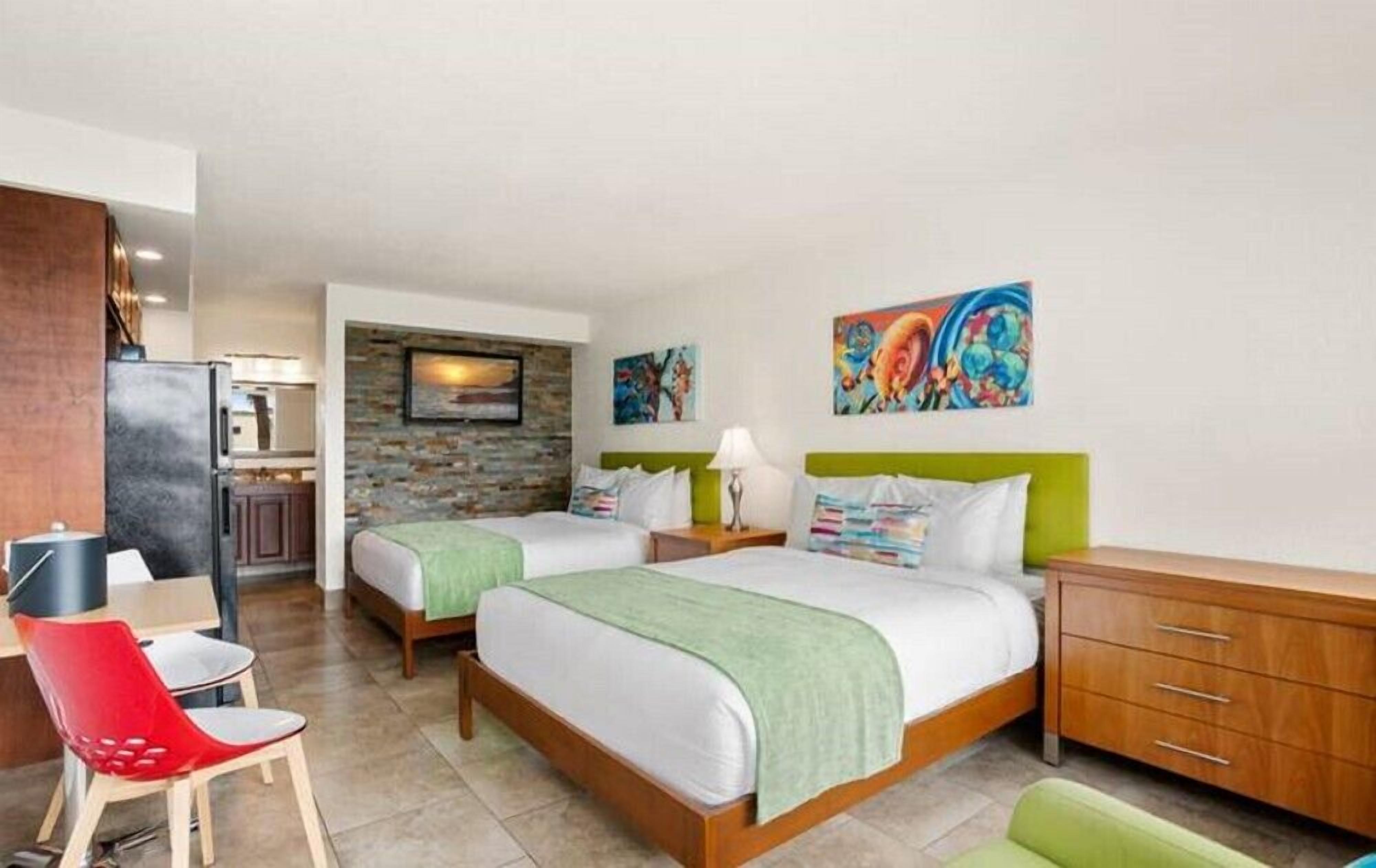 A hotel room with two beds, a desk, a chair, a kitchenette, and artwork on the walls.