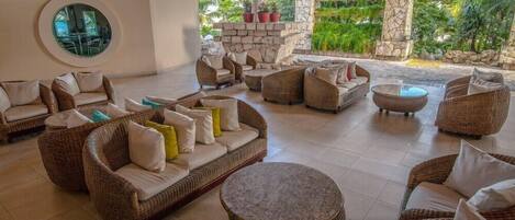 A spacious lounge area with wicker furniture, a round mirror, and a view of lush greenery.