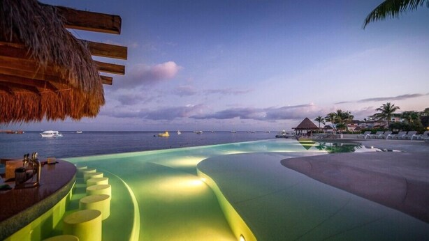 A pool area with a view of the ocean and boats, a thatched roof, and lounge chairs.