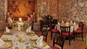 Un restaurante con paredes de piedra y un mural, mesas dispuestas con manteles blancos y detalles dorados, y sillas a rayas rojas y blancas.
