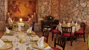 Ein Restaurant mit Steinsälen und einem Wandgemälde, Tische sind mit weißen Tischdecken und goldrandigen Tellern gedeckt, und es gibt rot gestreifte Stühle.