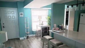 Eine Küche mit einem weißen Kühlschrank, einem Spülbecken und einem Fenster mit Jalousien. Eine Leiter lehnt an der Wand, und es gibt einen Tisch mit Hockern.