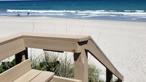 Eine Holzterrasse führt zu einem Strand mit klarem, blauem Wasser und Wellen.