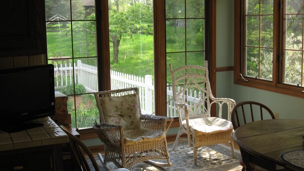 A sunlit room with a television, wicker chairs, a dining table, and large windows overlooking a white picket fence and greenery. - 3 Bedroom House in Rural Setting but close to Iowa City/Solon (Solon)