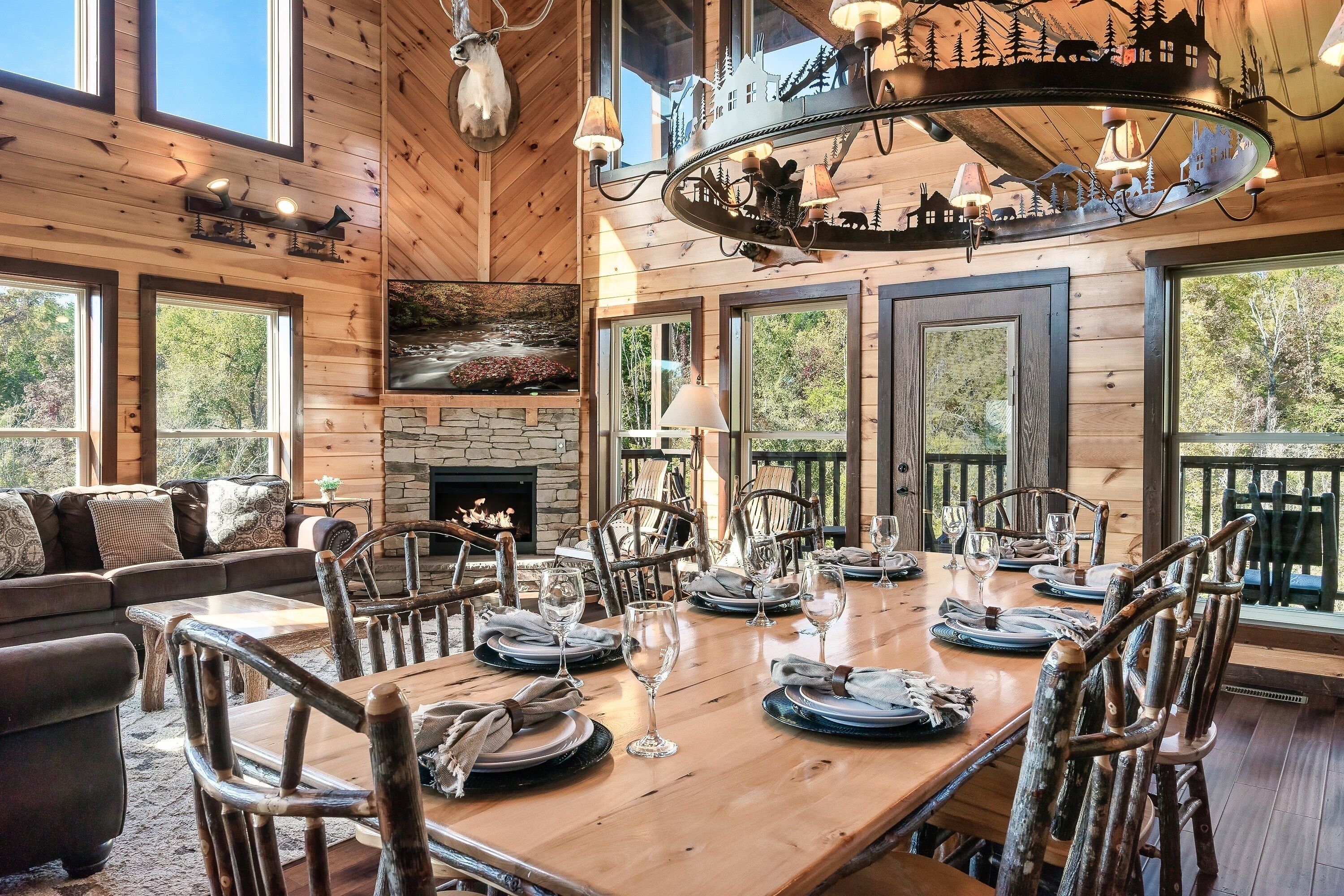 A wooden cabin interior with a dining table set for six, surrounded by rustic chairs, a fireplace, and large windows offering views of the outdoors.