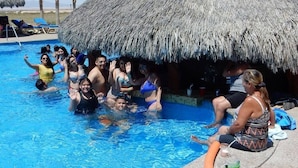 A group of people around a pool with a thatched-roof structure, palm trees, and lounge chairs in the background. - Shell Yeah! 🐚 🏝 Paradise 🦀 (Son)