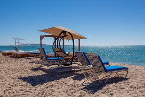 A beach with sun loungers and umbrellas, a clear sky, and a calm sea. - Shell Yeah! 🐚 🏝 Paradise 🦀 (Son)