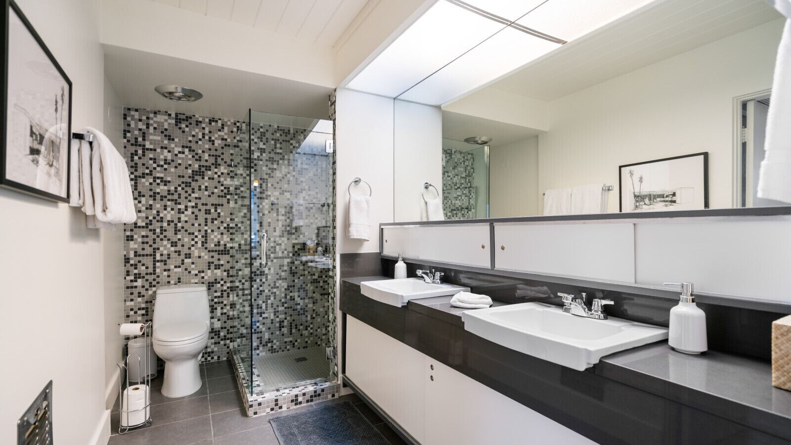 Un baño moderno con cabina de ducha de vidrio, inodoro blanco y un espejo grande sobre un mostrador negro con dos lavamanos.