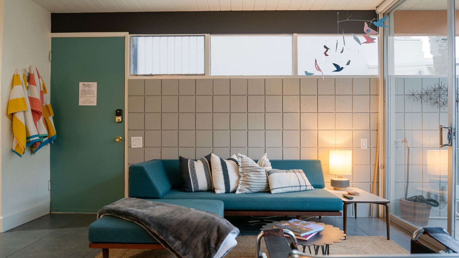 A living room with a teal sofa, patterned cushions, a small table with a lamp, and a window with blinds.