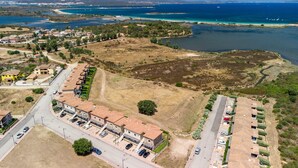 Vue aérienne d’un quartier résidentiel situé sur la côte, avec une rangée de maisons, un parking et, au loin, une vue sur la mer et les montagnes.
