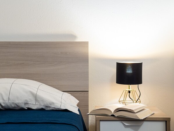 A neatly made bed with a white pillow and a blue blanket, a bedside table with an open book, and a modern lamp with a black shade. - Myrsine 7D - design flat, garden facing the bay (Olbia)