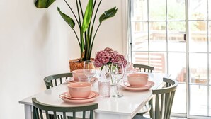 Une table à manger dressée avec des bols et des verres roses, entourée de chaises, avec une grande plante et une fenêtre en arrière-plan.