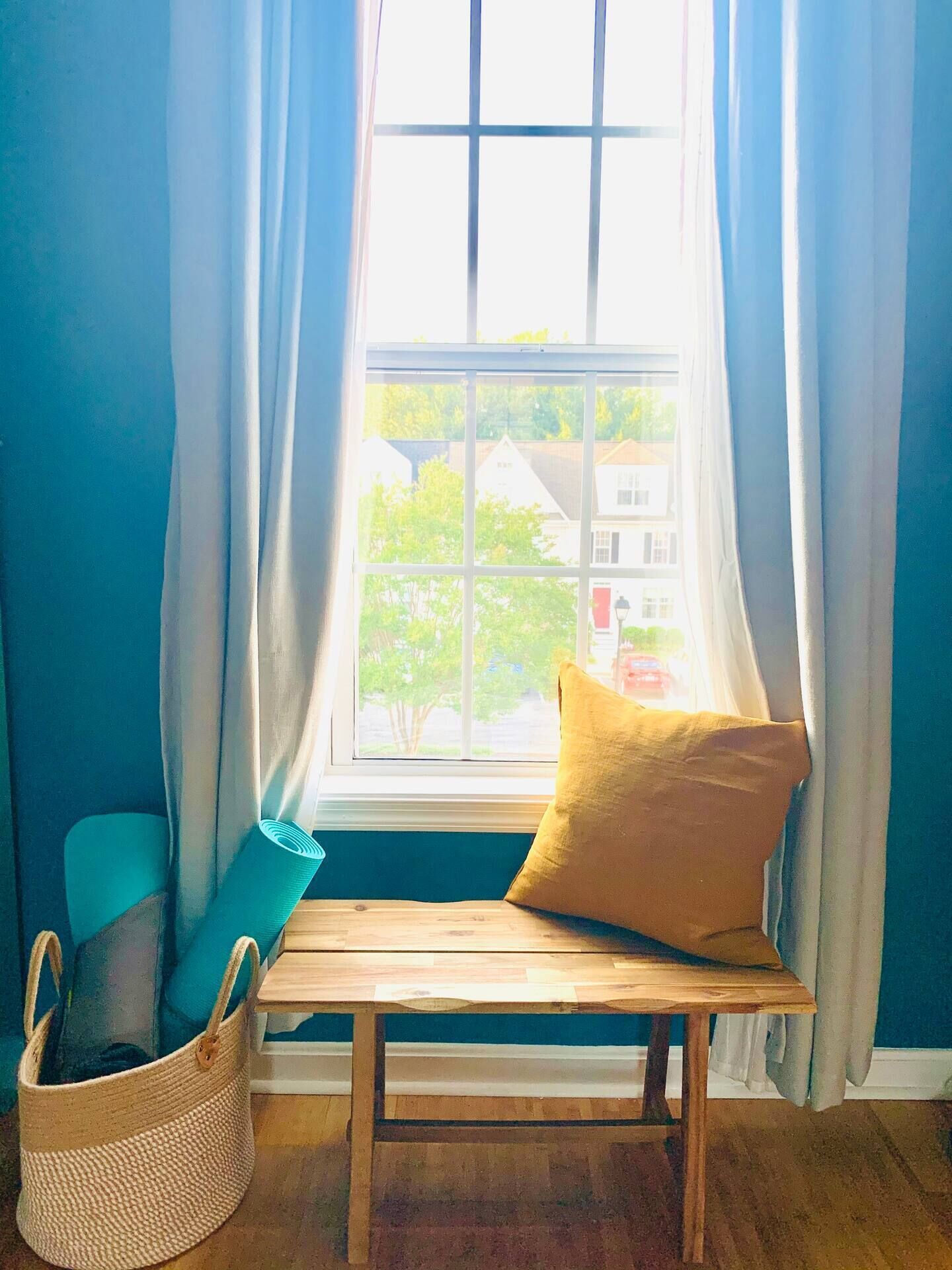 Una habitación con un banco de madera, un cojín amarillo, una cesta de mimbre y una ventana con cortinas.