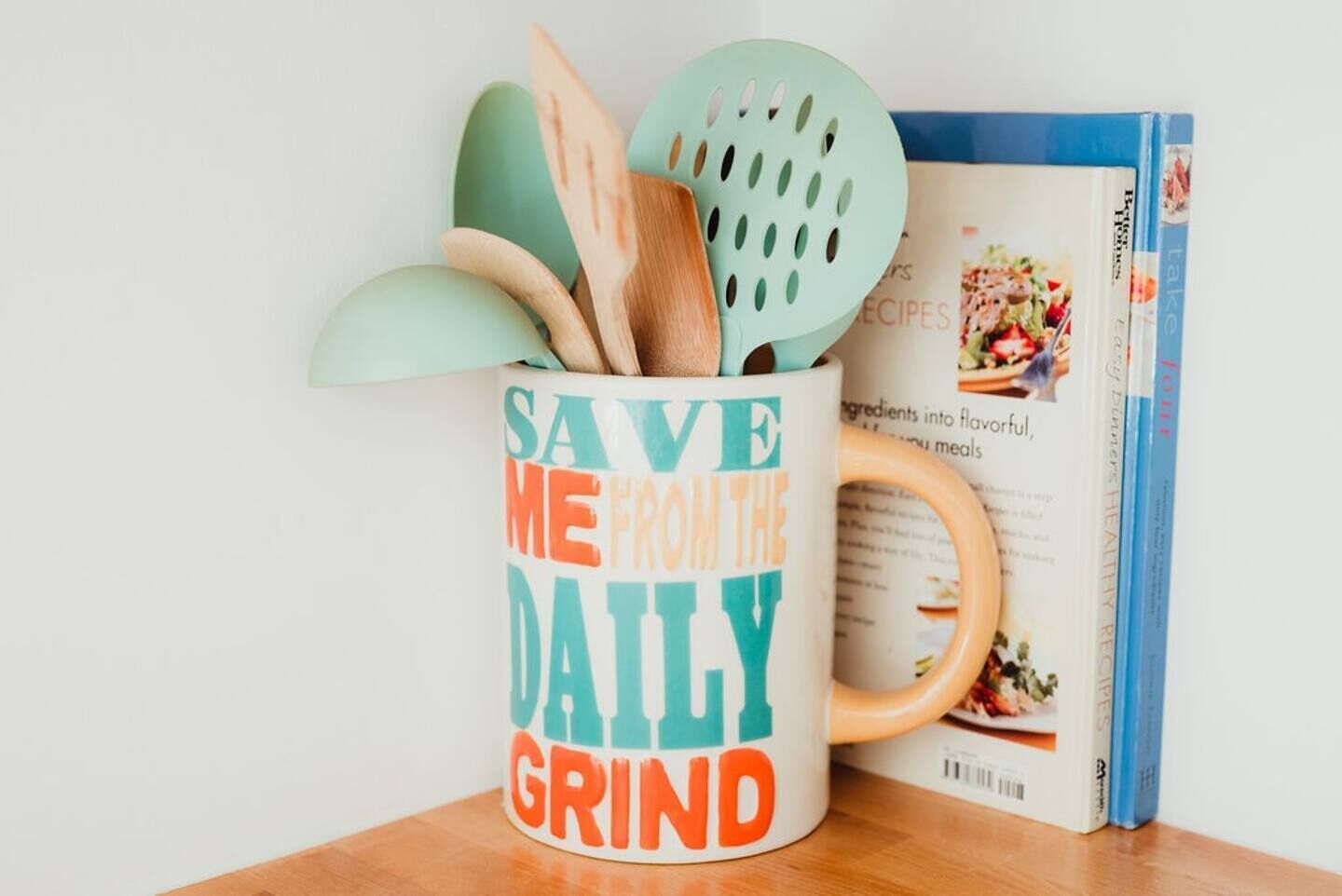 A kitchen utensil holder with a mug that reads "SAVE ME FROM THE DAILY GRIND" and a cookbook titled "Healthy Recipes for Busy Diners".