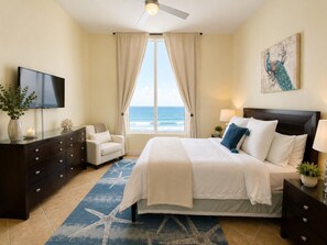 Quarto com cama, televisão, cômoda, cadeira e vista para o oceano.