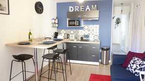 Eine kompakte Küche mit blauer Akzentwand, ein Essbereich mit Hockern und ein blaues Sofa mit gemusterten Kissen.