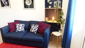 Ein Wohnzimmer mit einem dunkelblauen Sofa, roten und schwarzen Kissen mit Muster, einem kleinen weißen Beistelltisch mit Pflanze und einem roten Teppich.