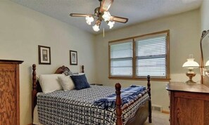 A bedroom with a bed, a ceiling fan, a window with blinds, a bedside table with a lamp, and framed pictures on the wall.