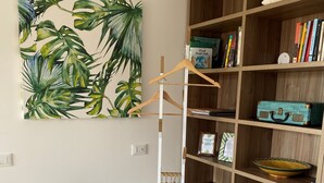Une pièce avec une bibliothèque en bois, un bureau avec une chaise blanche, un petit tabouret avec un siège à motifs floraux et un tableau représentant des feuilles tropicales accroché au mur.