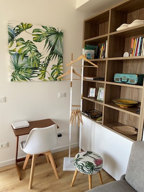 Een kamer met een houten boekenkast, een bureau met een witte stoel, een klein krukje met een bloemenmotief en een schilderij van tropische bladeren aan de muur.