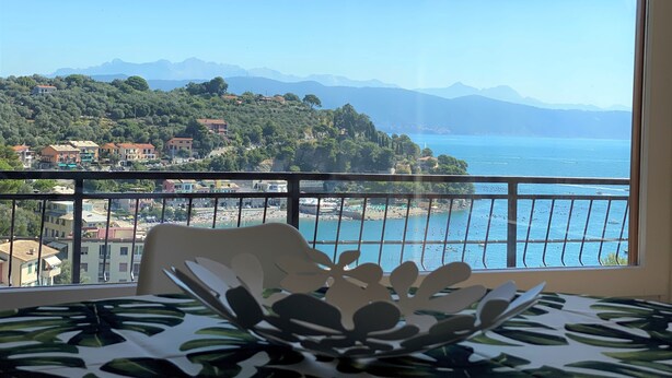 Un balcon donnant sur une ville côtière, une table à manger recouverte d’une nappe à motifs de feuilles et un vase décoratif.