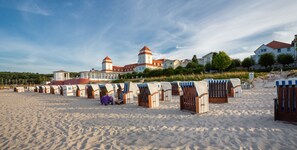 A beach with rows of beach huts numbered 9 and 92, a large building with red roofs in the background, and a clear sky.