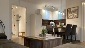 A modern living room with a sofa, a coffee table, and a dining area with chairs and a table. There is a kitchen area with cabinets and a microwave. A glass shower door is visible in the background. - Exclusive vacation apartment, Residence Margarete, close to the beach, underground parking, sauna (Binz)