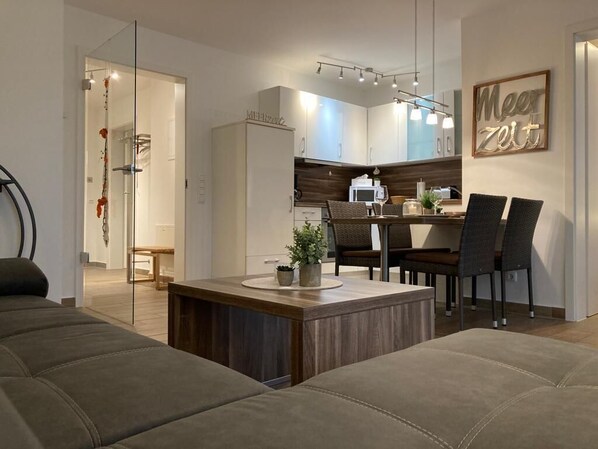 A modern living room with a sofa, a coffee table, and a dining area with chairs and a table. There is a kitchen area with cabinets and a microwave. A glass shower door is visible in the background.