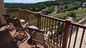 A wooden deck with two chairs and a table, overlooking a residential area with houses and a forested hill. - Luxury Ski-in/Ski-out 2 BR, Soaring Eagle 207 (Snowshoe)