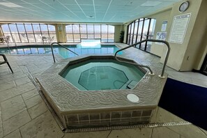 Une piscine intérieure dotée d’un jacuzzi, entourée d’un sol carrelé et de grandes fenêtres offrant une vue sur l’extérieur.