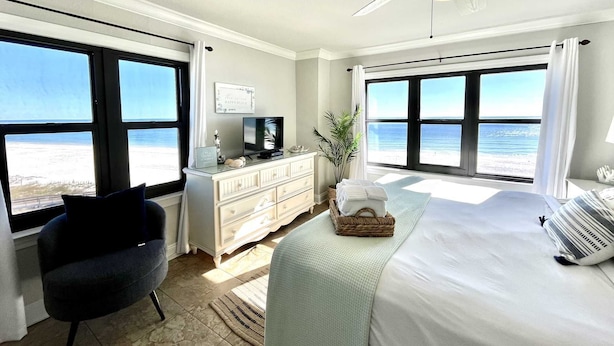 Een slaapkamer met een bed, een bureau, een stoel, een plafondventilator en grote ramen met uitzicht op het strand.