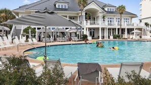 Una gran piscina con sillones y sombrillas, rodeada por un edificio de varios pisos con balcones.